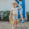 Child wearing a blue and white striped swim nappy outdoors with a wicker basket and another child in the background.