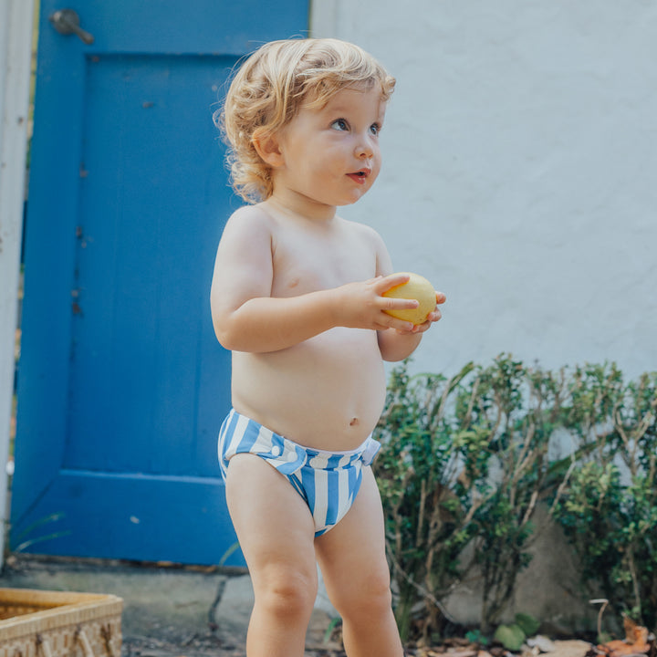 Child in crywolf blue and white striped swim nappy holding an apple outdoors.