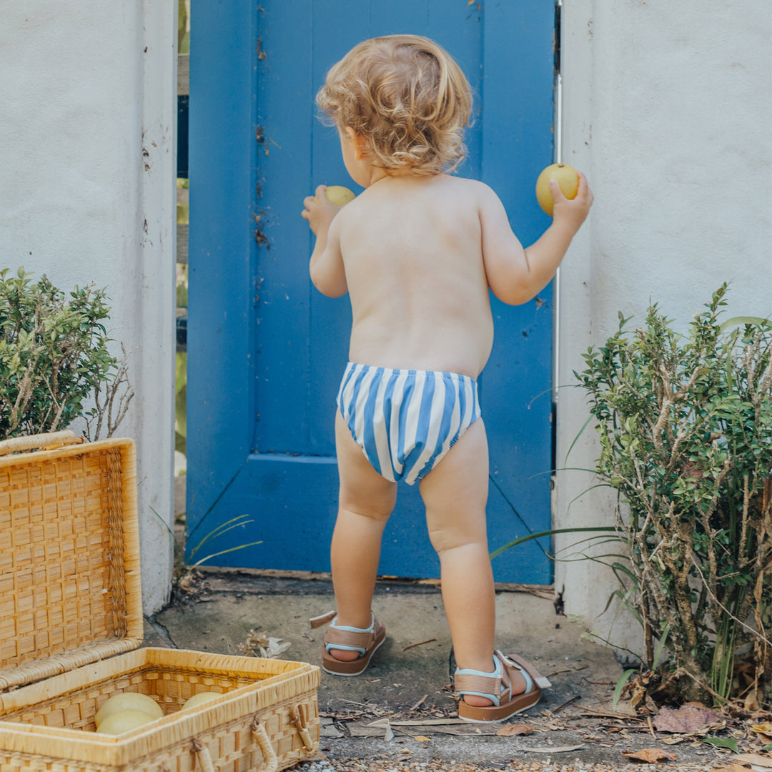 Child in crywolf striped swim diaper holding apples outdoors near a blue door.