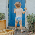 Child in crywolf striped swim diaper holding apples outdoors near a blue door.