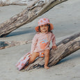 Child in a pink crywolf outfit and hat sitting on a log at the beach