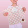 Child holding a pink and white checkered bag with a visible brand label against a pink background