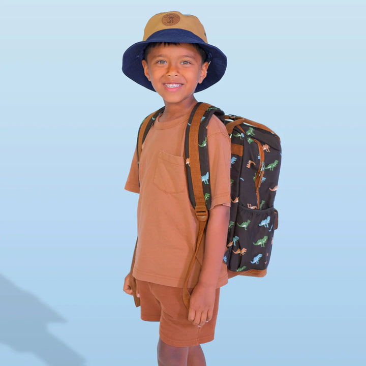 Child wearing a brown shirt, shorts, and a blue hat with a dinosaur-patterned backpack on a light blue background