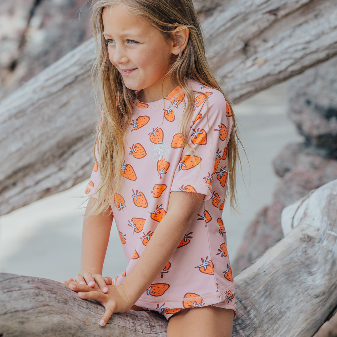 Young girl in a crywolf pink rash set with orange patterns sitting on driftwood.