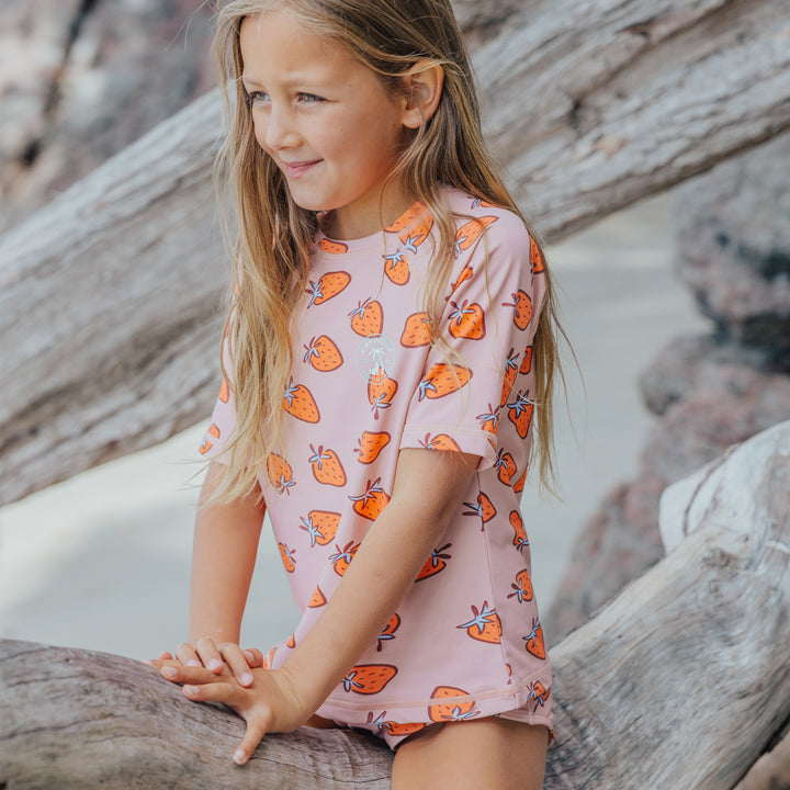 Young girl in a crywolf pink rash set with orange patterns sitting on driftwood.