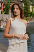 Woman wearing a light-colored one-shoulder top and pants by a body of water with buildings in the background.