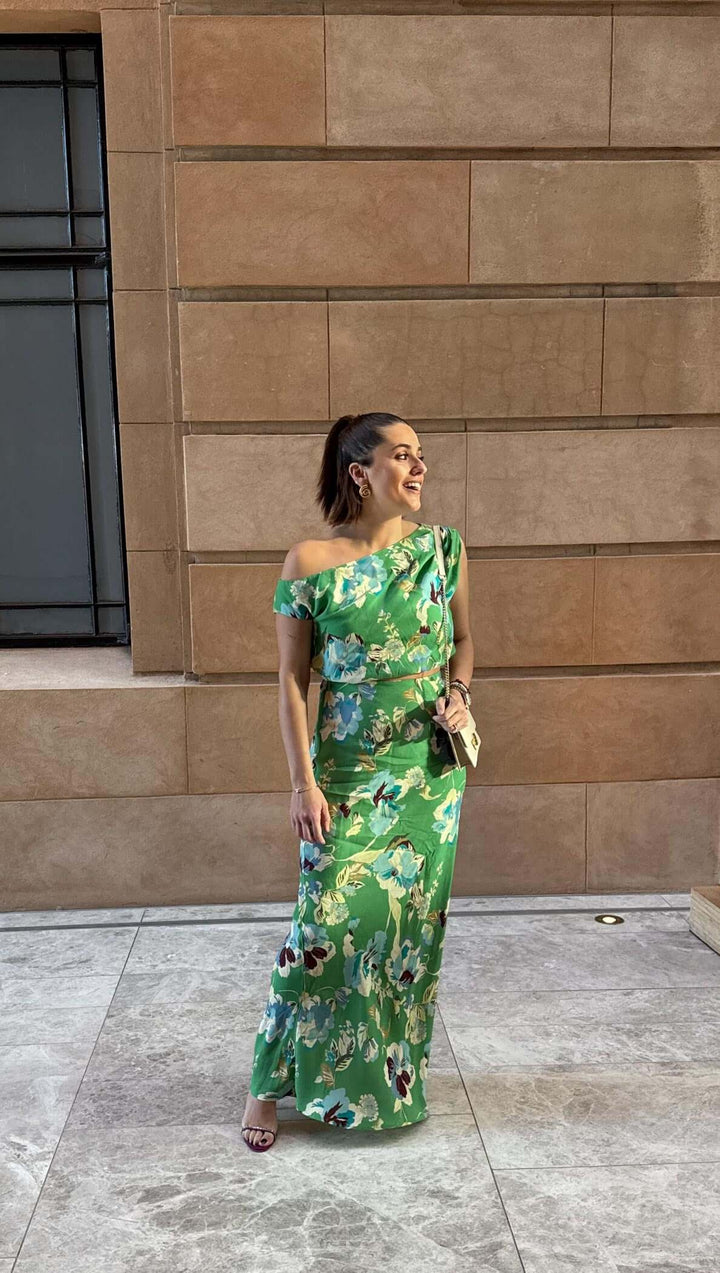 Woman in a green floral dress standing against a beige brick wall.