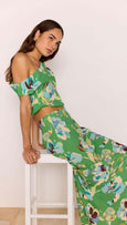 Woman wearing a green floral outfit sitting on a white stool against a white wall.