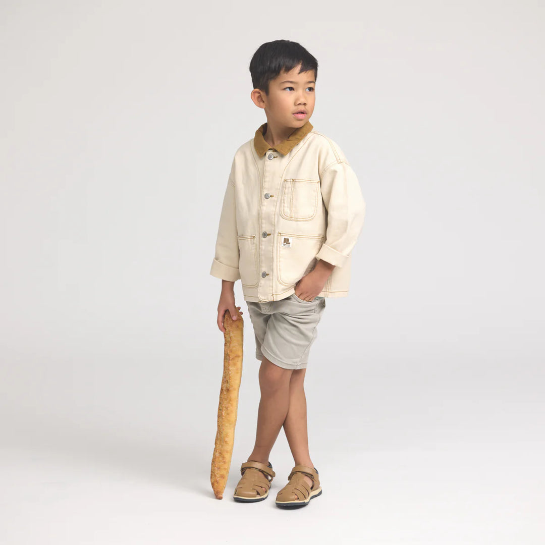 Child wearing a beige jacket and shorts holding a wooden walking stick on a white background