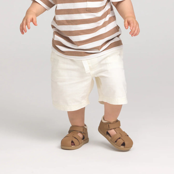 Child wearing a striped shirt and white pants with brown sandals on a plain background