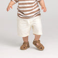 Child wearing a striped shirt and white pants with brown sandals on a plain background