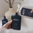 Person pouring body wash from a white bottle into a black bottle labeled 'alive'.