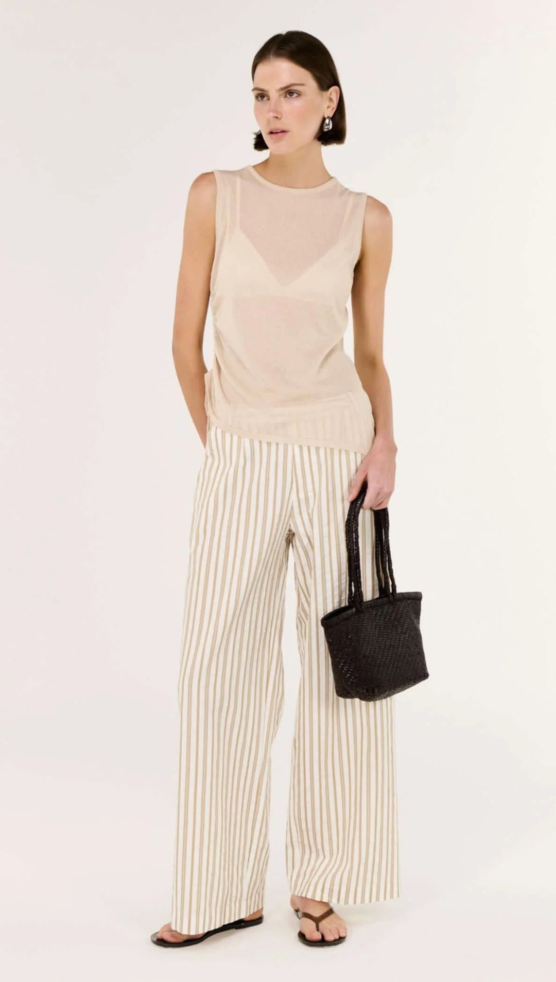 Woman wearing a beige sleeveless top and wide-leg pants, holding a black handbag on a white background
