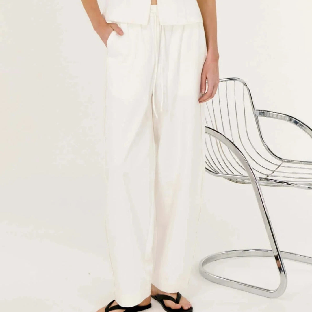 Woman wearing a white sleeveless top and pants with a chair in the background.