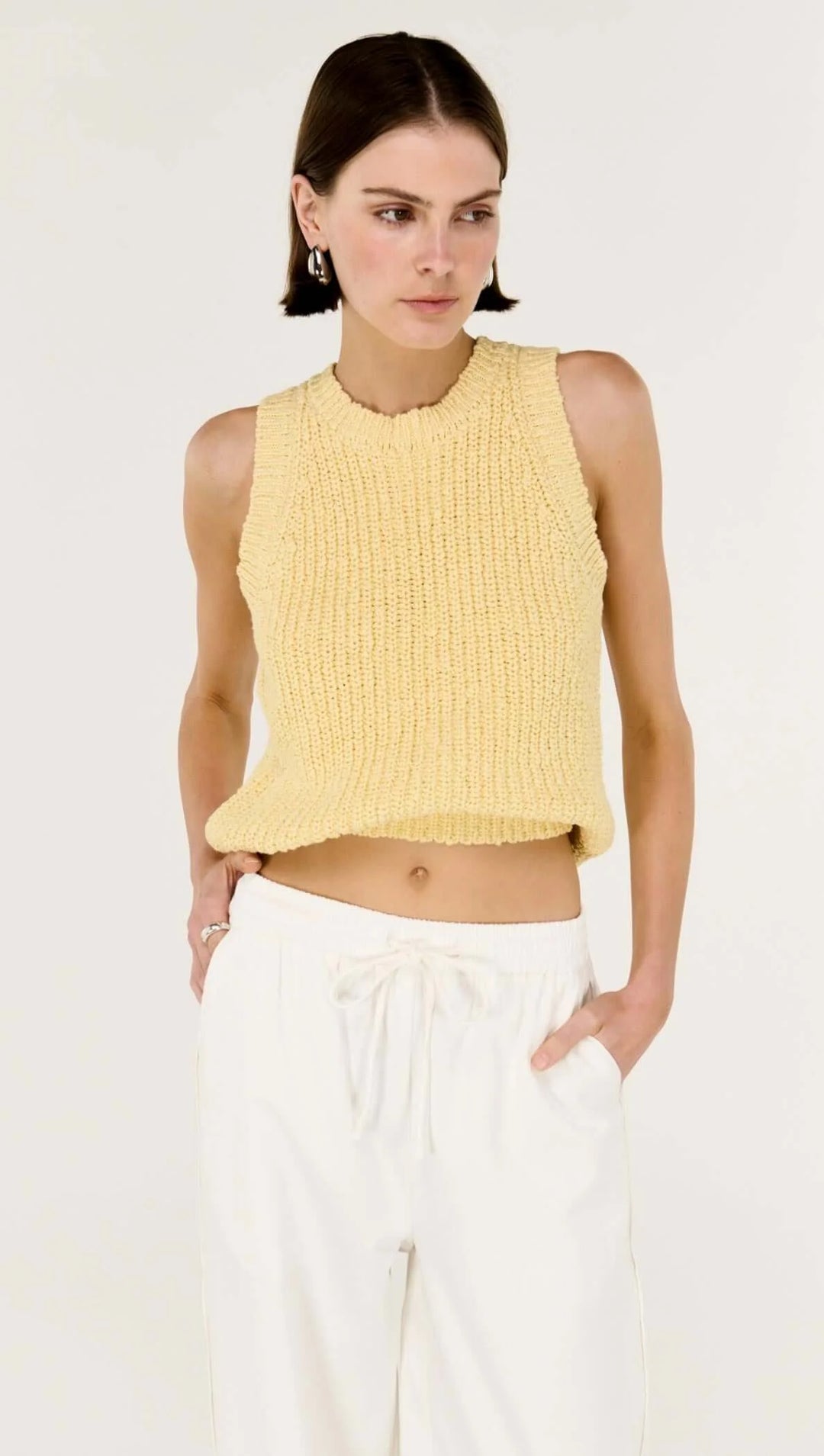 Woman wearing a yellow sleeveless top and white pants on a white background
