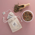 Tea packaging including a drawstring bag, tea leaves, and a wooden scoop on a pink background.