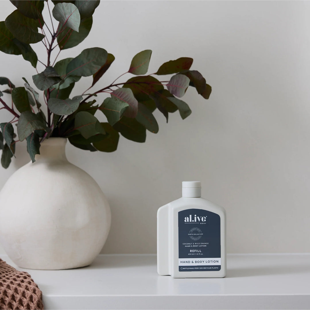 White bottle of al.ive hand and body lotion on a white surface with a vase and plant in the background.