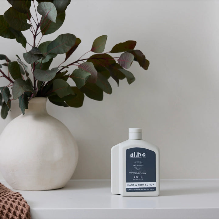 White bottle of al.ive hand and body lotion on a white surface with a vase and plant in the background.