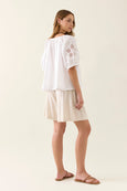 Woman wearing a white isle of mine blouse with embroidery and beige shorts on a plain background