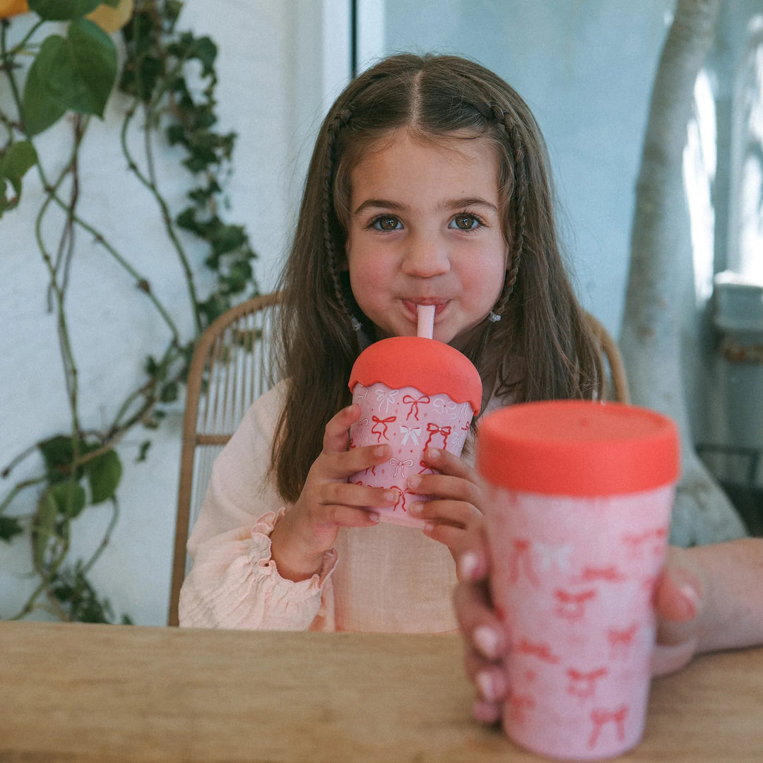 Young girl drinking from a pink tumbler with a red lid in a casual setting.