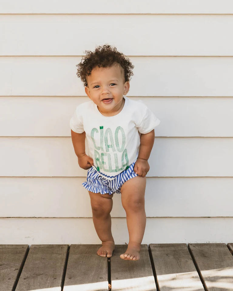Child wearing a white shirt with green text and striped diaper standing on a wooden deck.
