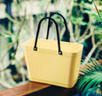 Yellow tote bag with black handles on a wooden surface with greenery in the background