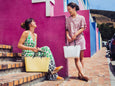 Two women sitting on a colorful street with a pink wall and blue sky.