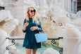 Woman in teal dress holding a blue handbag in front of a fountain
