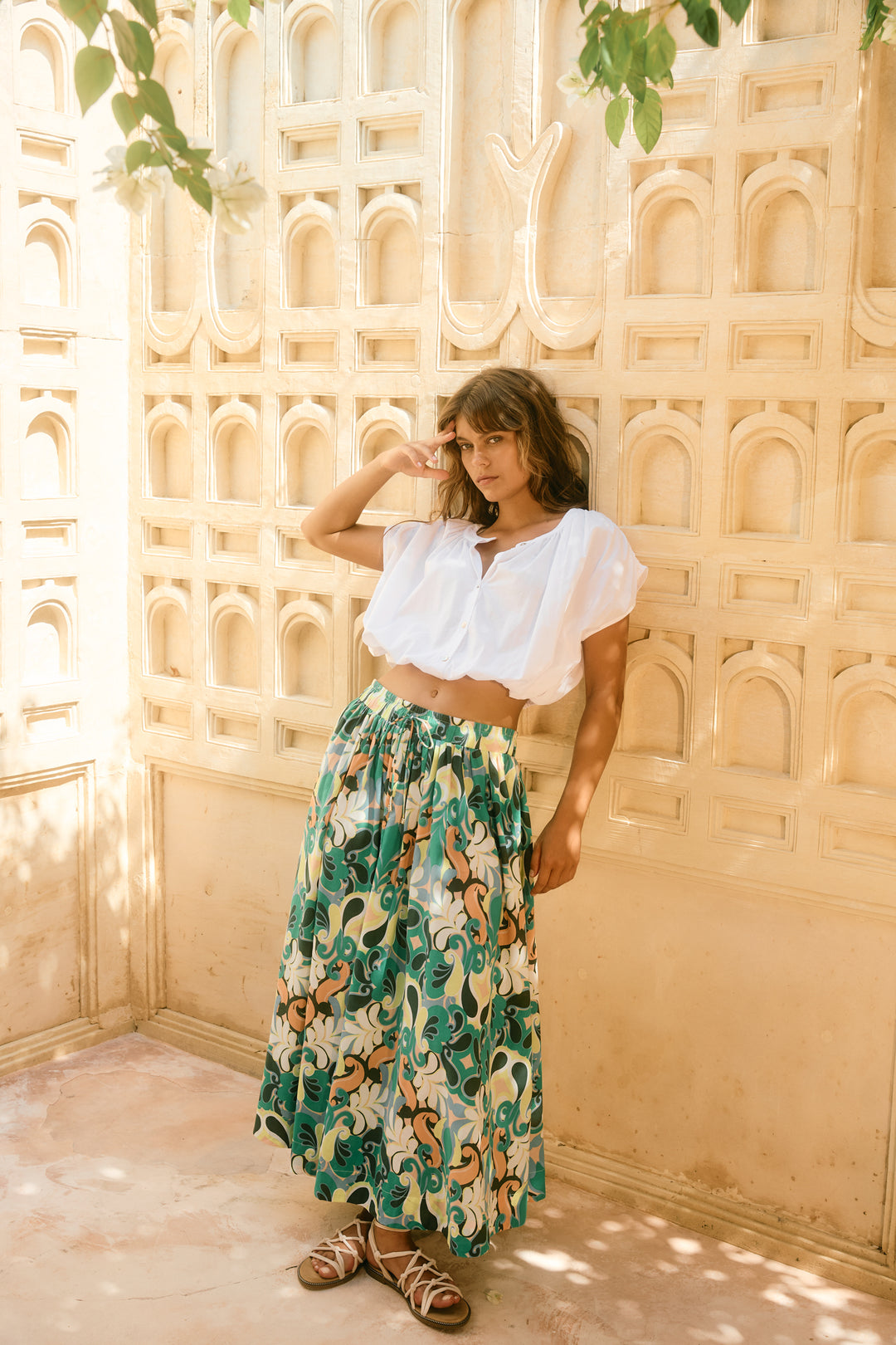 Woman in a white top and floral isle of mine skirt standing against a decorative wall.