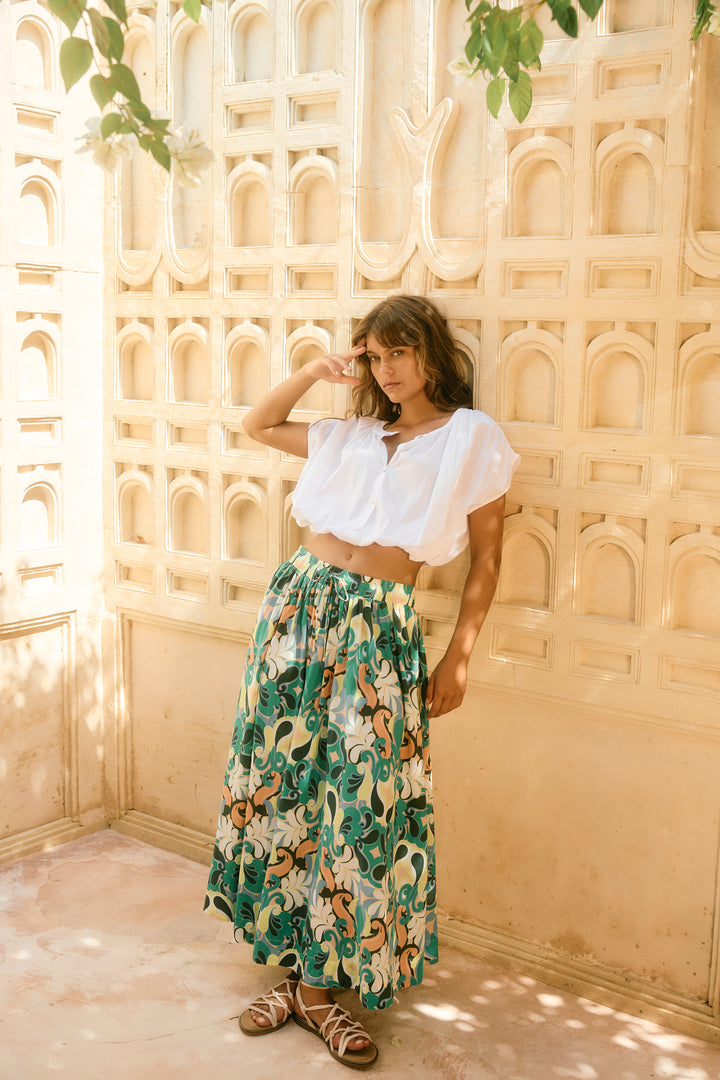 Woman in a white top and floral isle of mine skirt standing against a decorative wall.