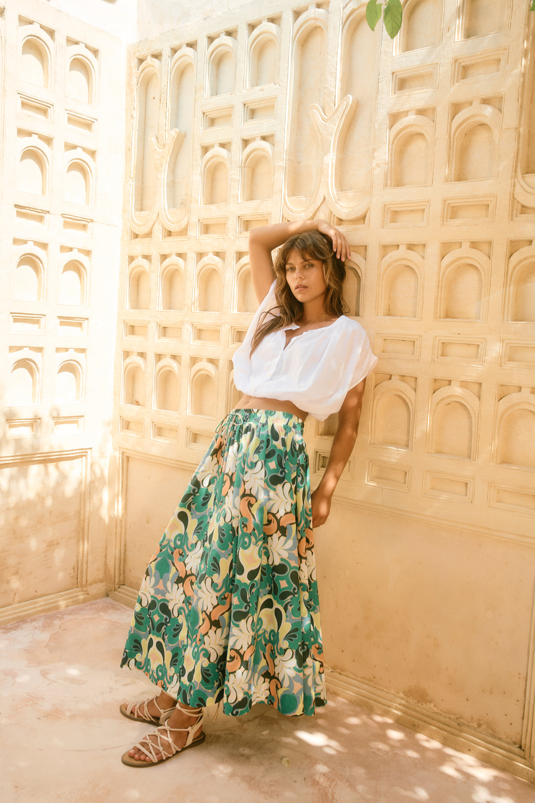 Woman in a white top and floral isle of mine skirt standing against a decorative wall.