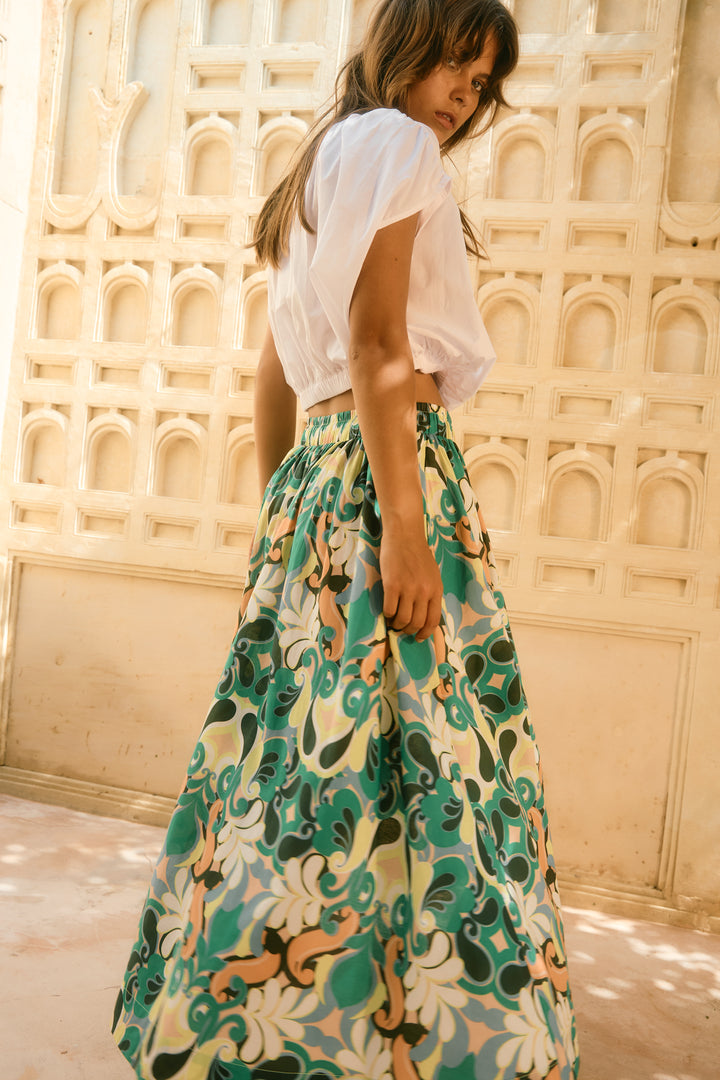 Woman wearing a white top and isle of mine floral skirt against a beige wall with arches.