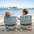 Two children sitting on checkered beach chairs facing the ocean with a city skyline in the background.