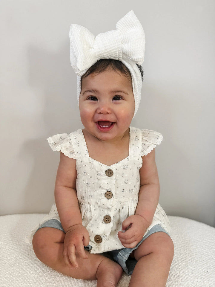 Baby wearing a white floral top with buttons and a large white bow headband.
