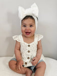 Baby wearing a white floral top with buttons and a large white bow headband.