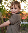 Child in a brown jubee & co shirt standing outdoors with trees and sunlight in the background