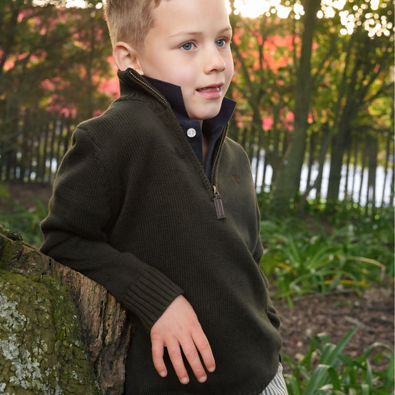 Young boy in a dark jubee & co sweater standing outdoors with trees and grass in the background