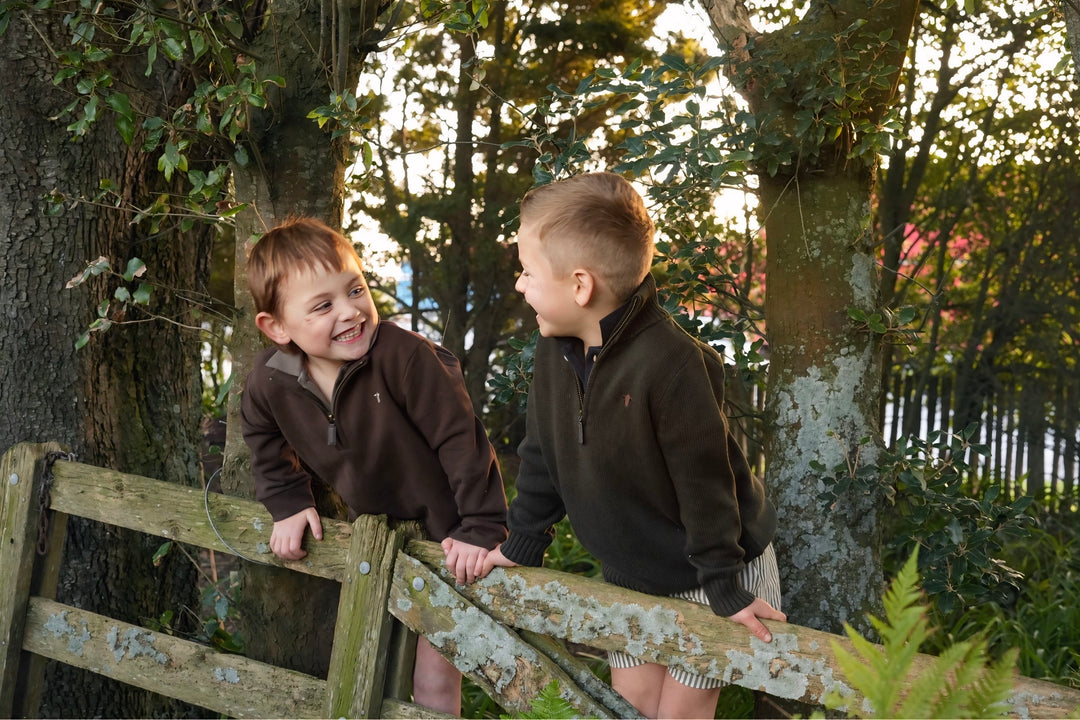Two children in matching jackets standing on a wooden fence with trees in the background