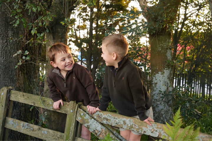 Two children in matching jackets standing on a wooden fence with trees in the background