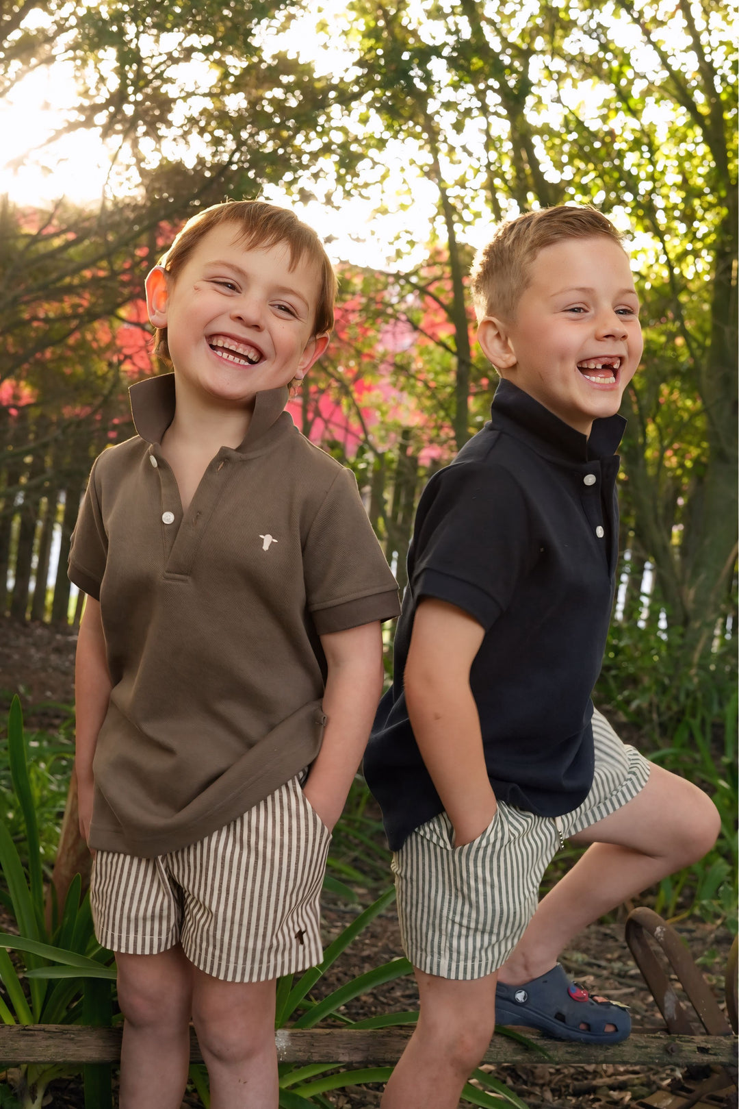 Two boys in jubee & co polo shirts and shorts standing outdoors with trees and flowers in the background.