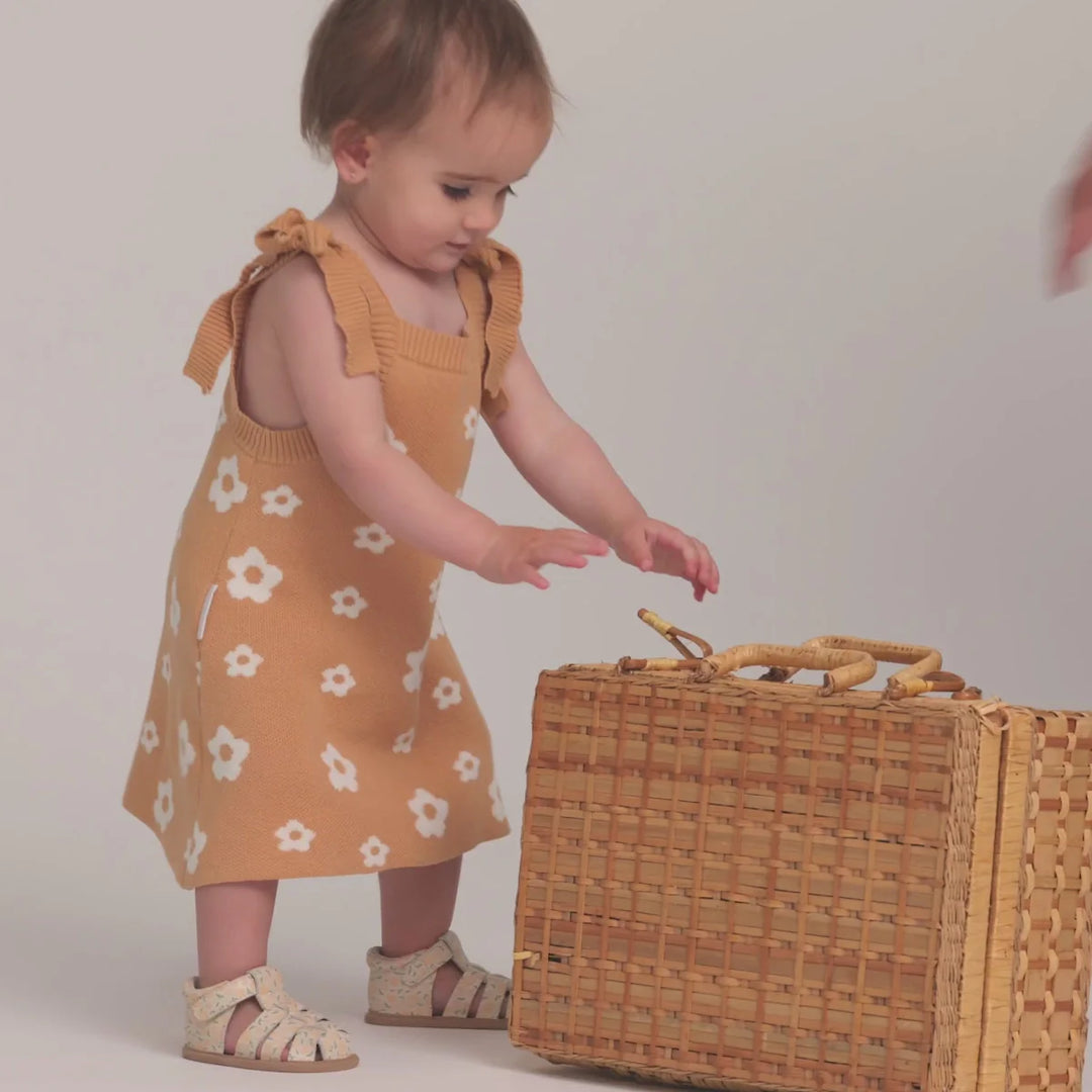 Child in a yellow dress with floral pattern interacting with a wicker basket on a plain background