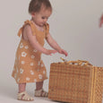 Child in a yellow dress with floral pattern interacting with a wicker basket on a plain background