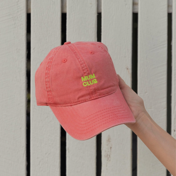 Pink cap with 'Mum Club' text held against a white wooden background