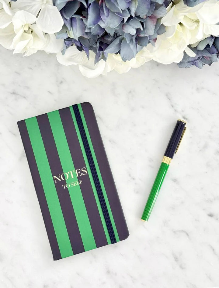 Notebook with green and black striped cover and pen on a marble surface with flowers.
