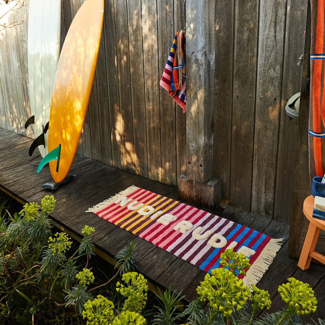 Colorful striped doormat with 'Happy Birthday' on a wooden deck next to a surfboard and towel.