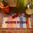 Colorful doormat with 'NUDIE RUDIE' text on a brick floor, surrounded by plants and a small table.