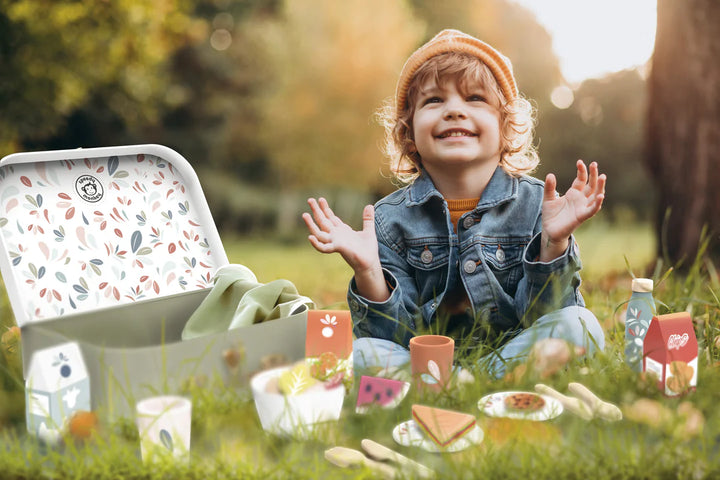 Child sitting outdoors with a picnic setup, including a lunch box and food items.