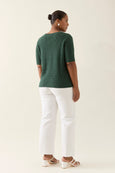 Woman wearing a green knitted isle of mine top and white pants on a plain background