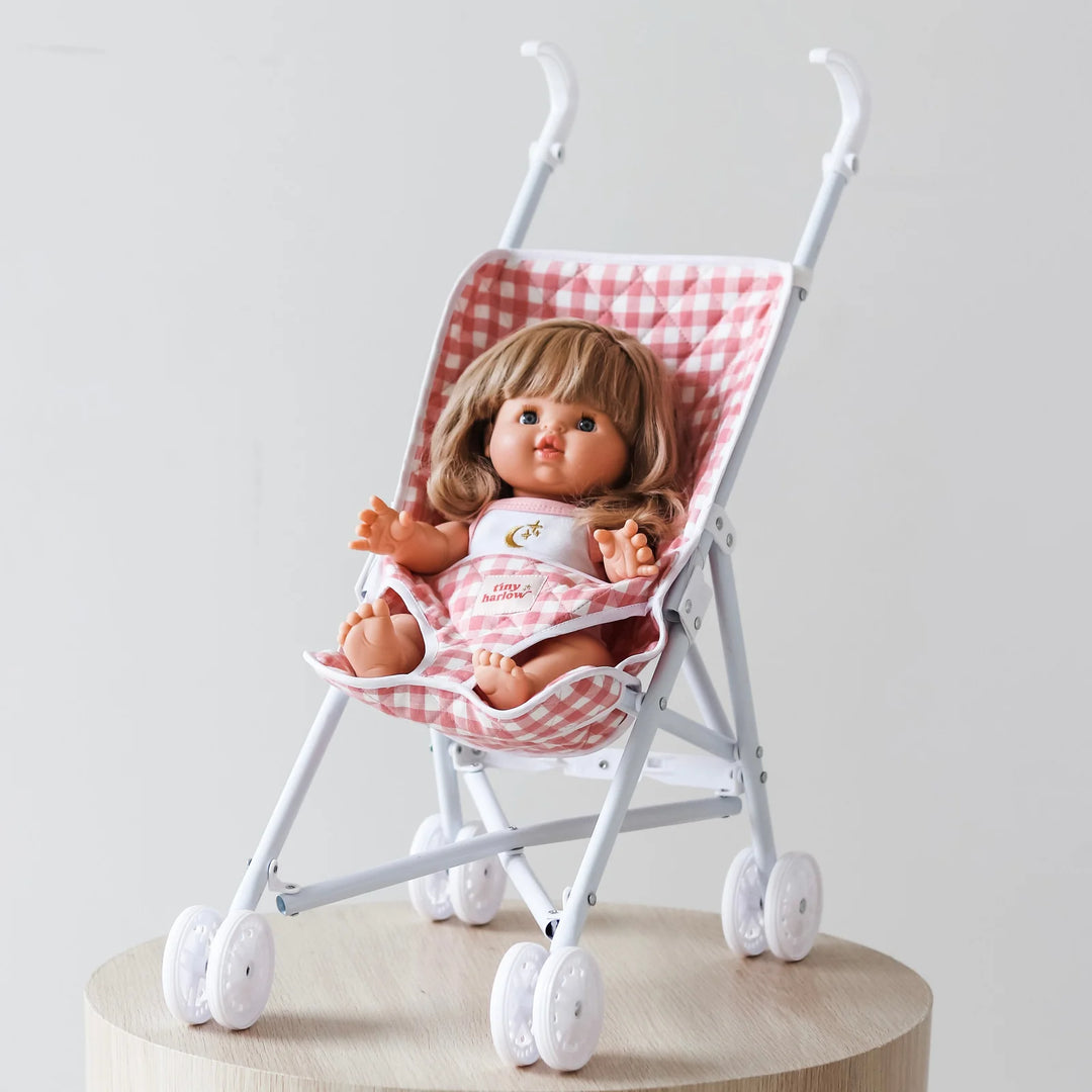 Doll in a small Tiny Harlow stroller with a checkered pink and white cover on a light gray background