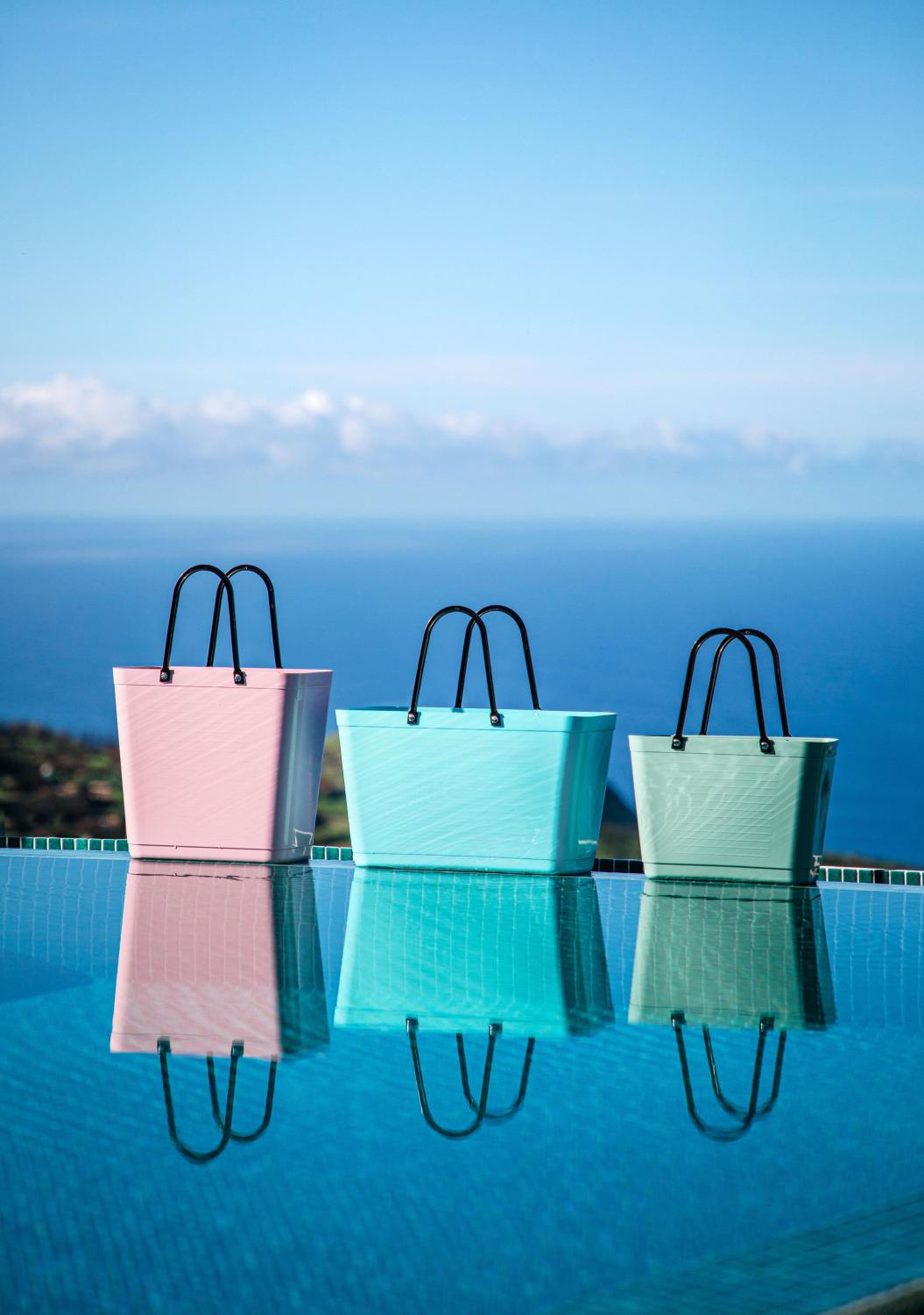 Three bags (pink, blue, green) reflected in water with a blue sky background